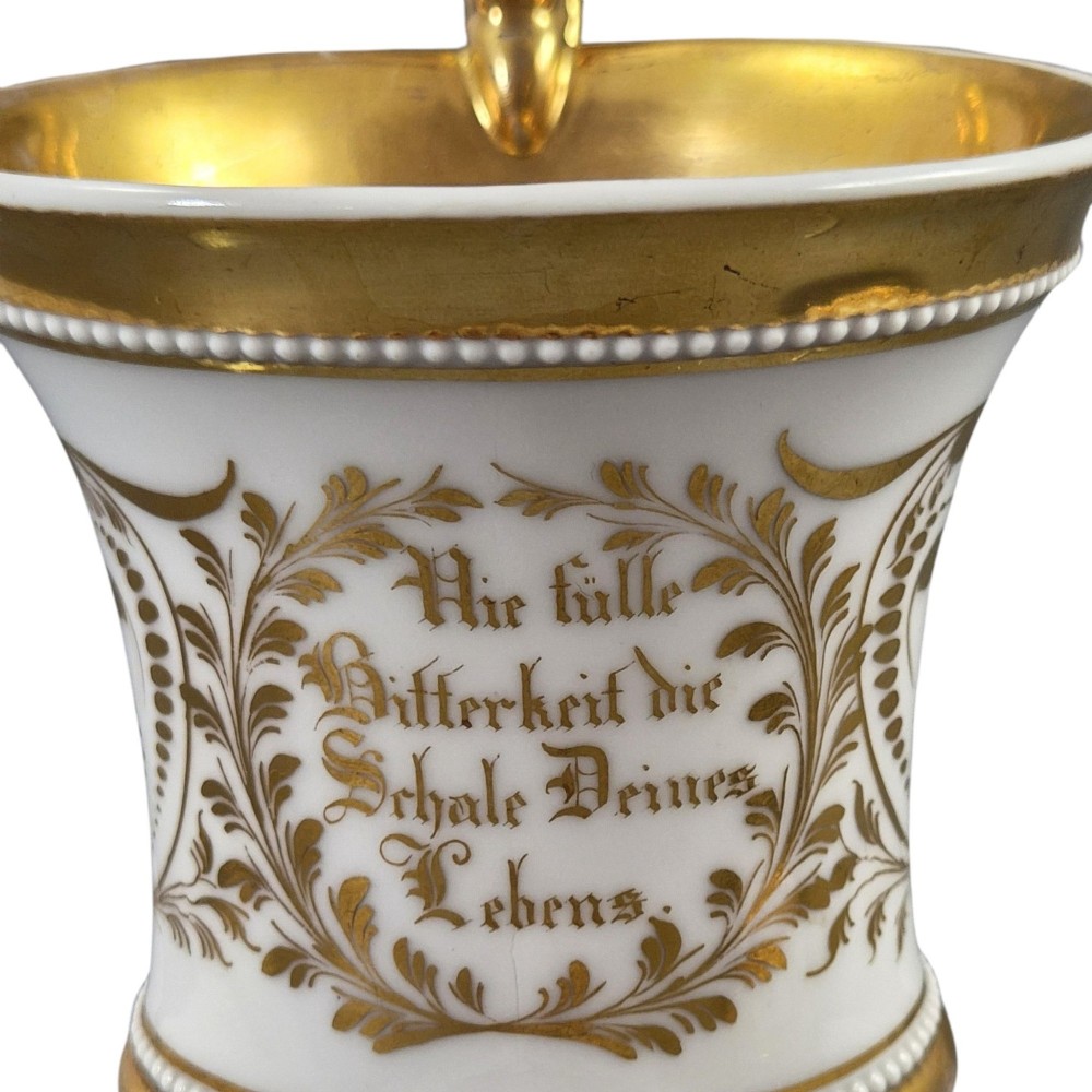 An antique white and gilt porcelain presentation cup with an elevated loop handle sitting on a matching gilded saucer. View X