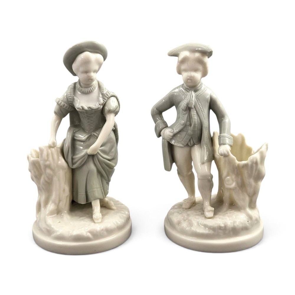A pair of late Victorian grey and cream glazed porcelain figural salts depicting a man and woman beside hollow tree stump vessels.