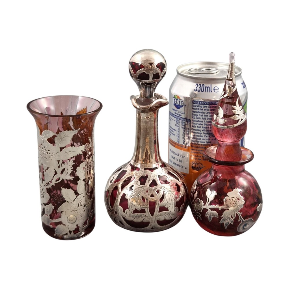A three-piece ruby red glass vanity set featuring floral sterling silver overlay details across two stoppered bottles and a cylindrical beaker. View XI