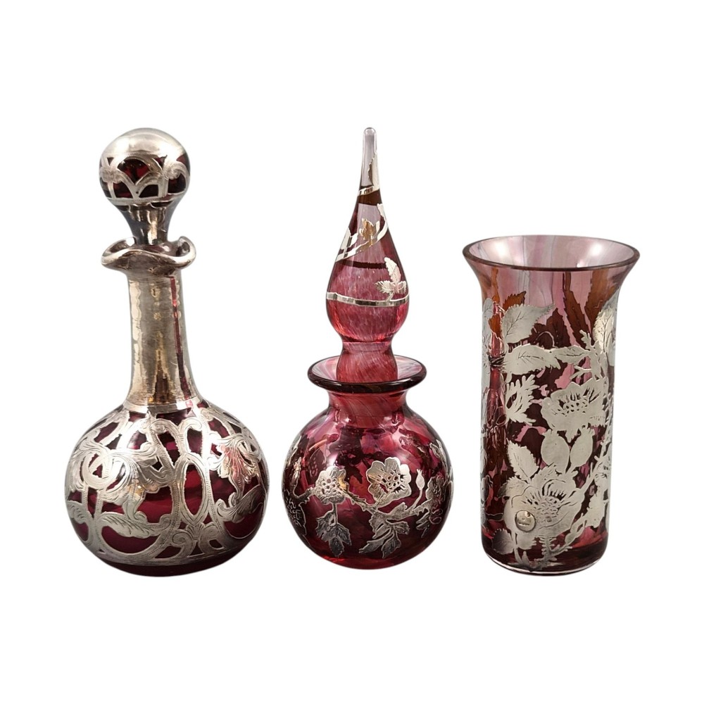 A three-piece ruby red glass vanity set featuring floral sterling silver overlay details across two stoppered bottles and a cylindrical beaker. View XII