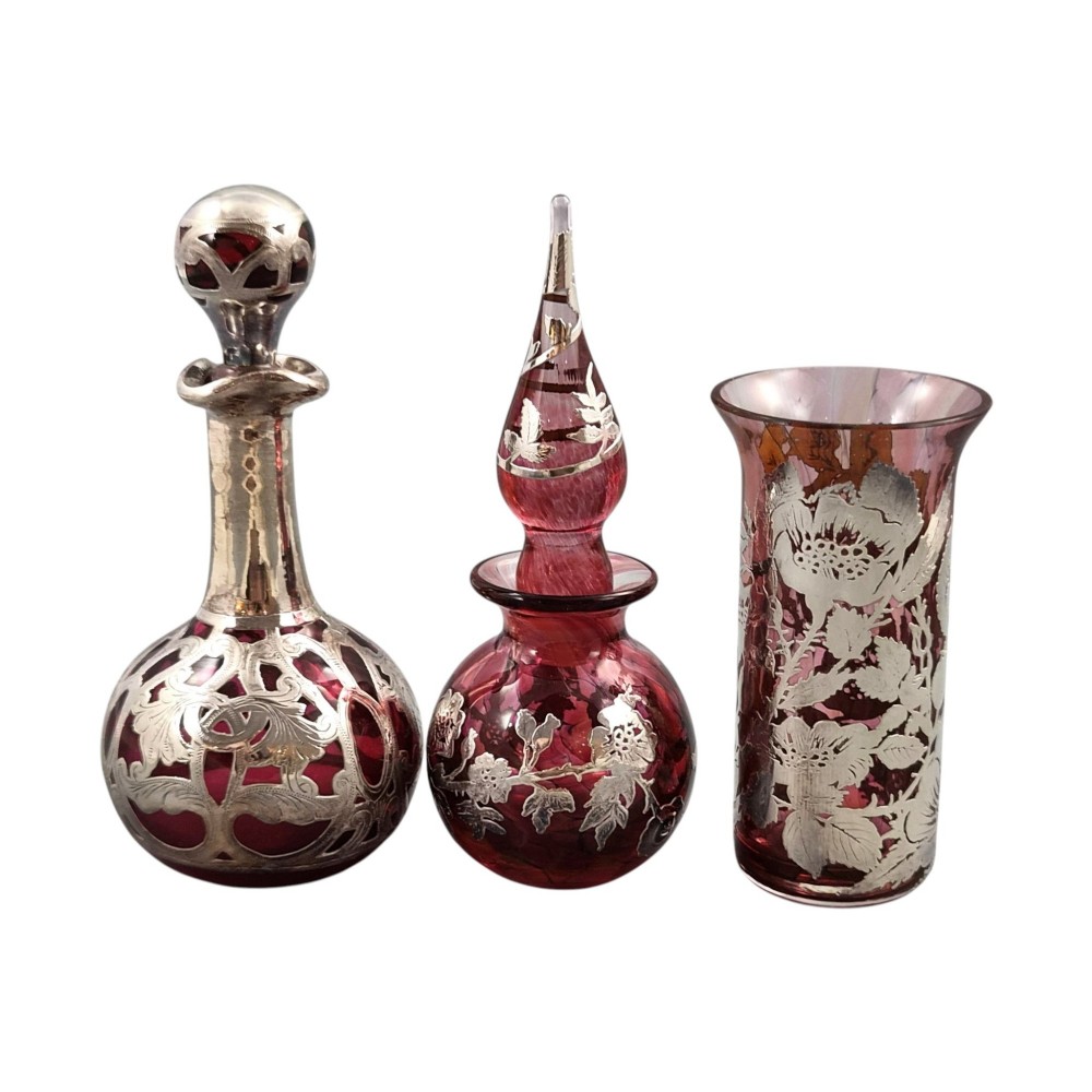 A three-piece ruby red glass vanity set featuring floral sterling silver overlay details across two stoppered bottles and a cylindrical beaker. View XIII