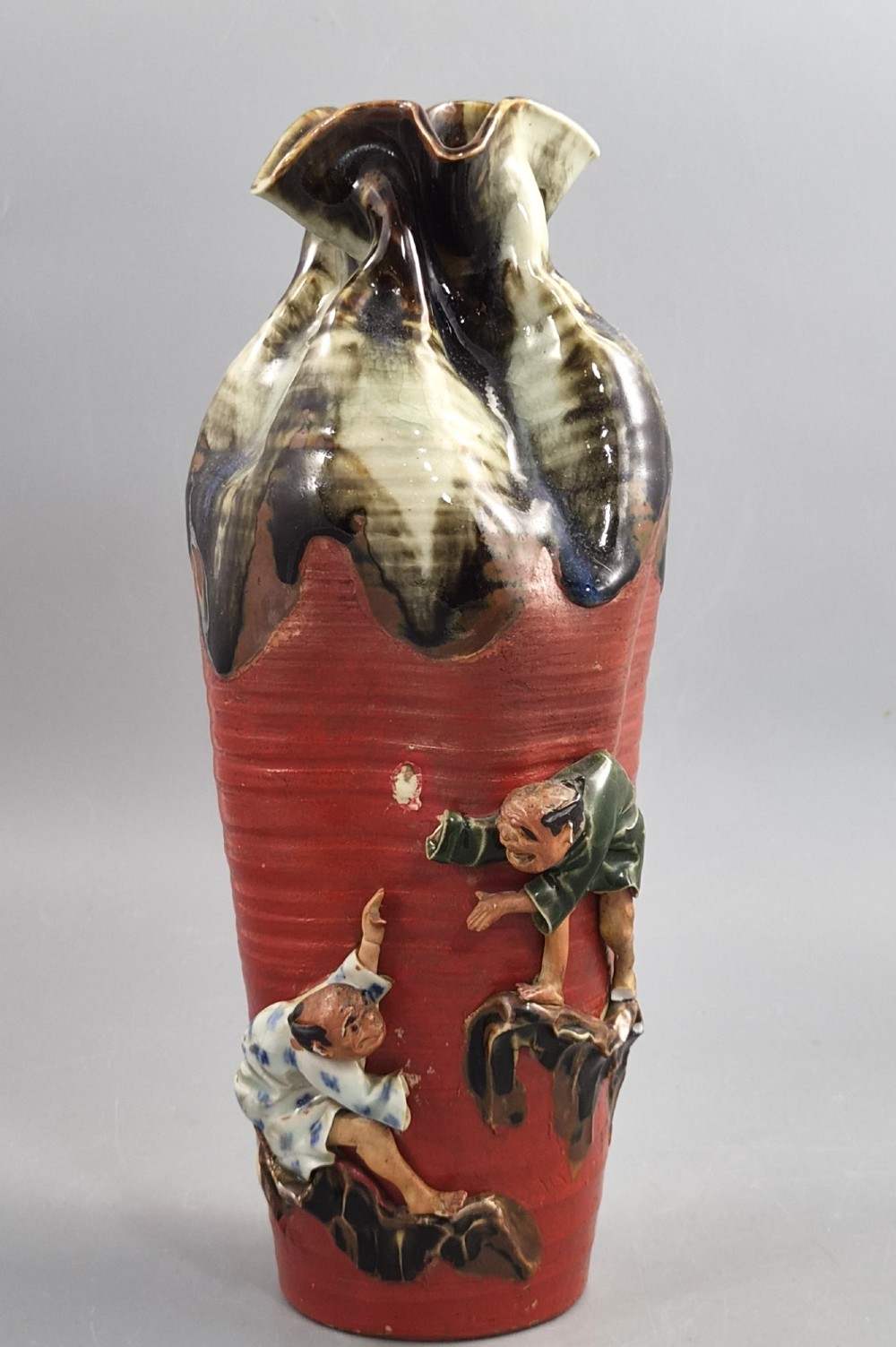 A tall Japanese Sumida Gawa ceramic vase featuring a ribbed red body, applied figures of climbing men, and a heavy flambe glaze around a pinched rim.