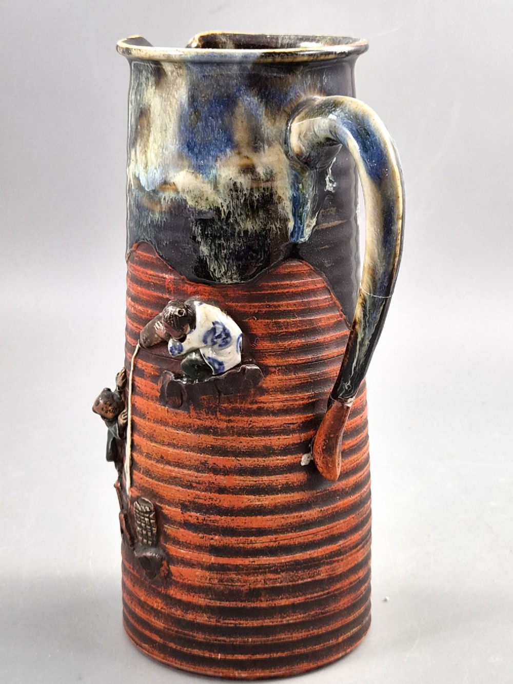 A tall Japanese Sumida Gawa ceramic pitcher featuring a ribbed terracotta body with relief figures pouring sake and a dark drip glaze near the lip. View II