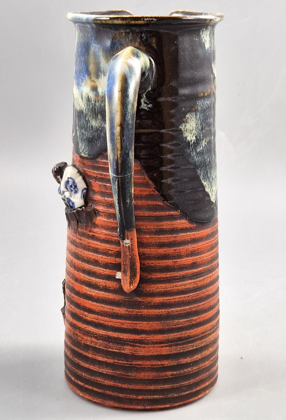 A tall Japanese Sumida Gawa ceramic pitcher featuring a ribbed terracotta body with relief figures pouring sake and a dark drip glaze near the lip. View III