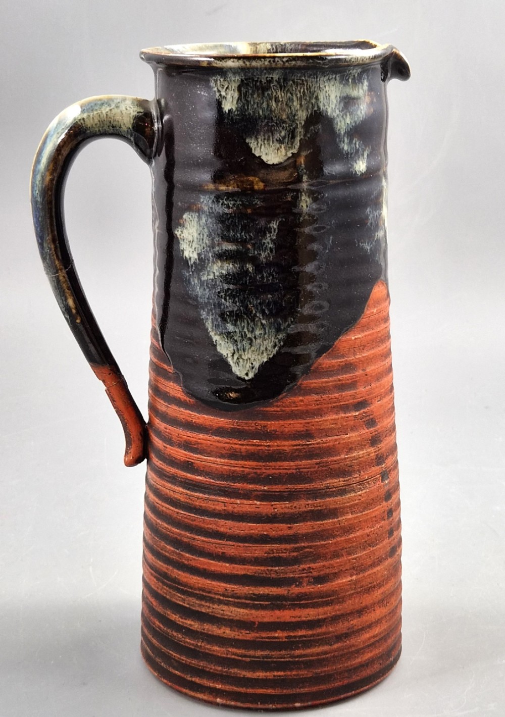 A tall Japanese Sumida Gawa ceramic pitcher featuring a ribbed terracotta body with relief figures pouring sake and a dark drip glaze near the lip. View IV