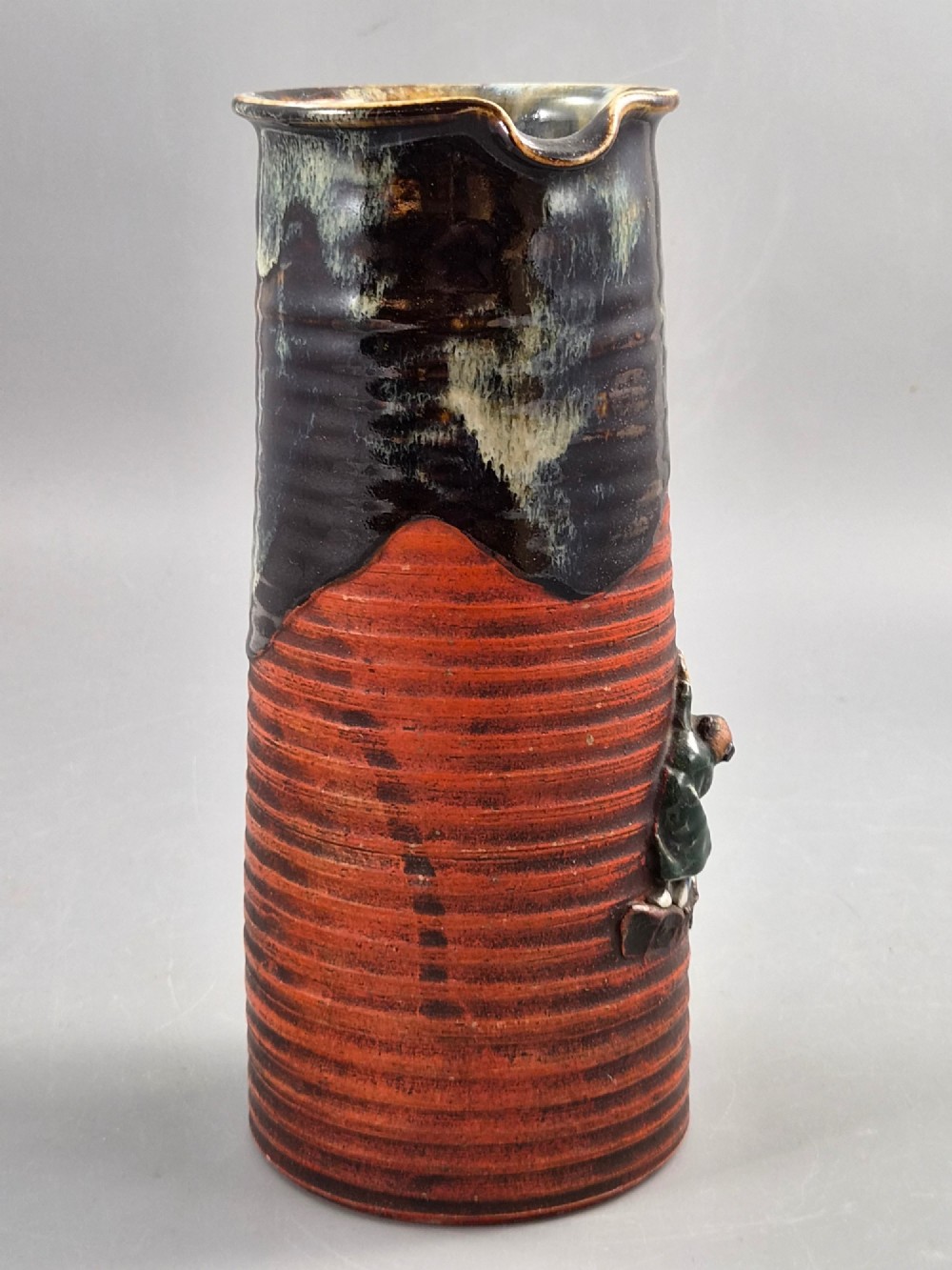 A tall Japanese Sumida Gawa ceramic pitcher featuring a ribbed terracotta body with relief figures pouring sake and a dark drip glaze near the lip. View V