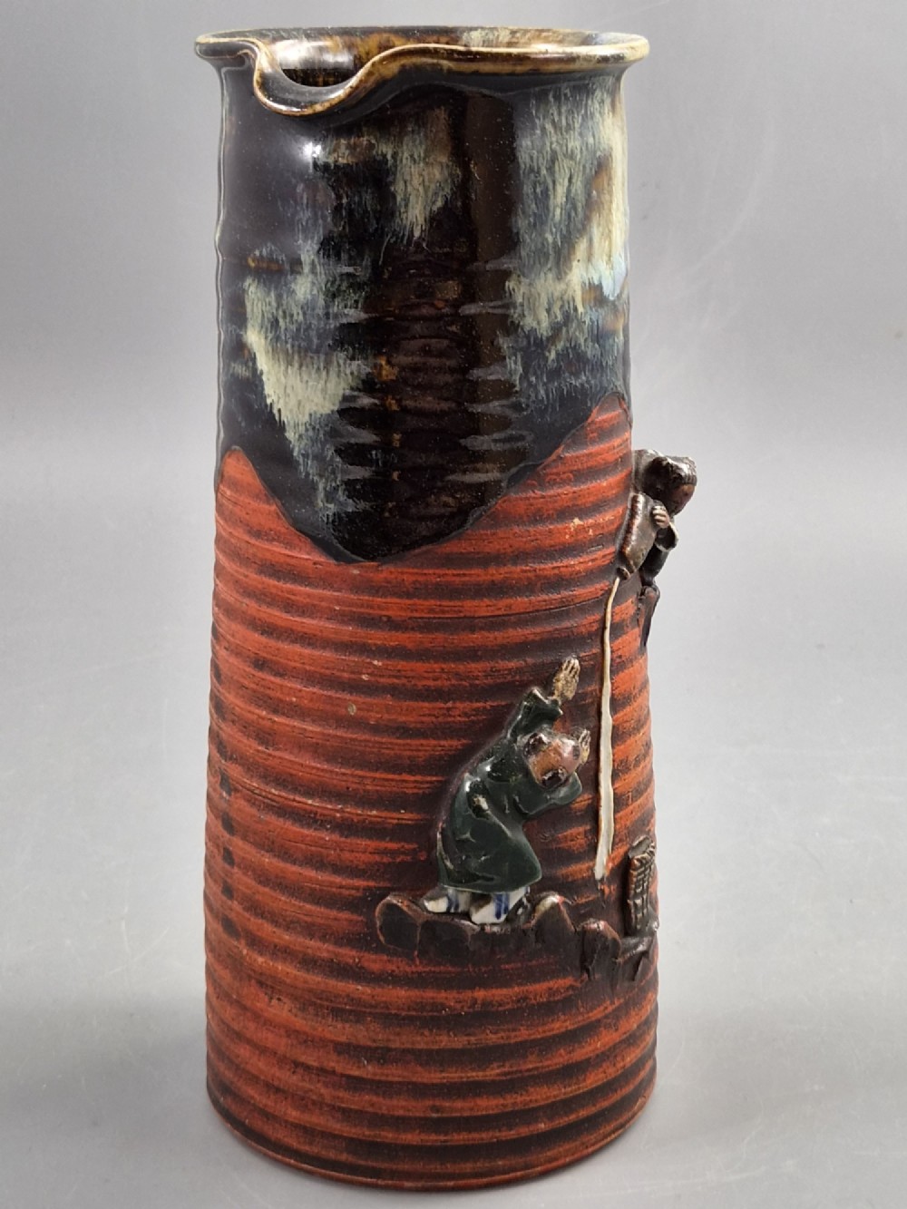 A tall Japanese Sumida Gawa ceramic pitcher featuring a ribbed terracotta body with relief figures pouring sake and a dark drip glaze near the lip. View VI