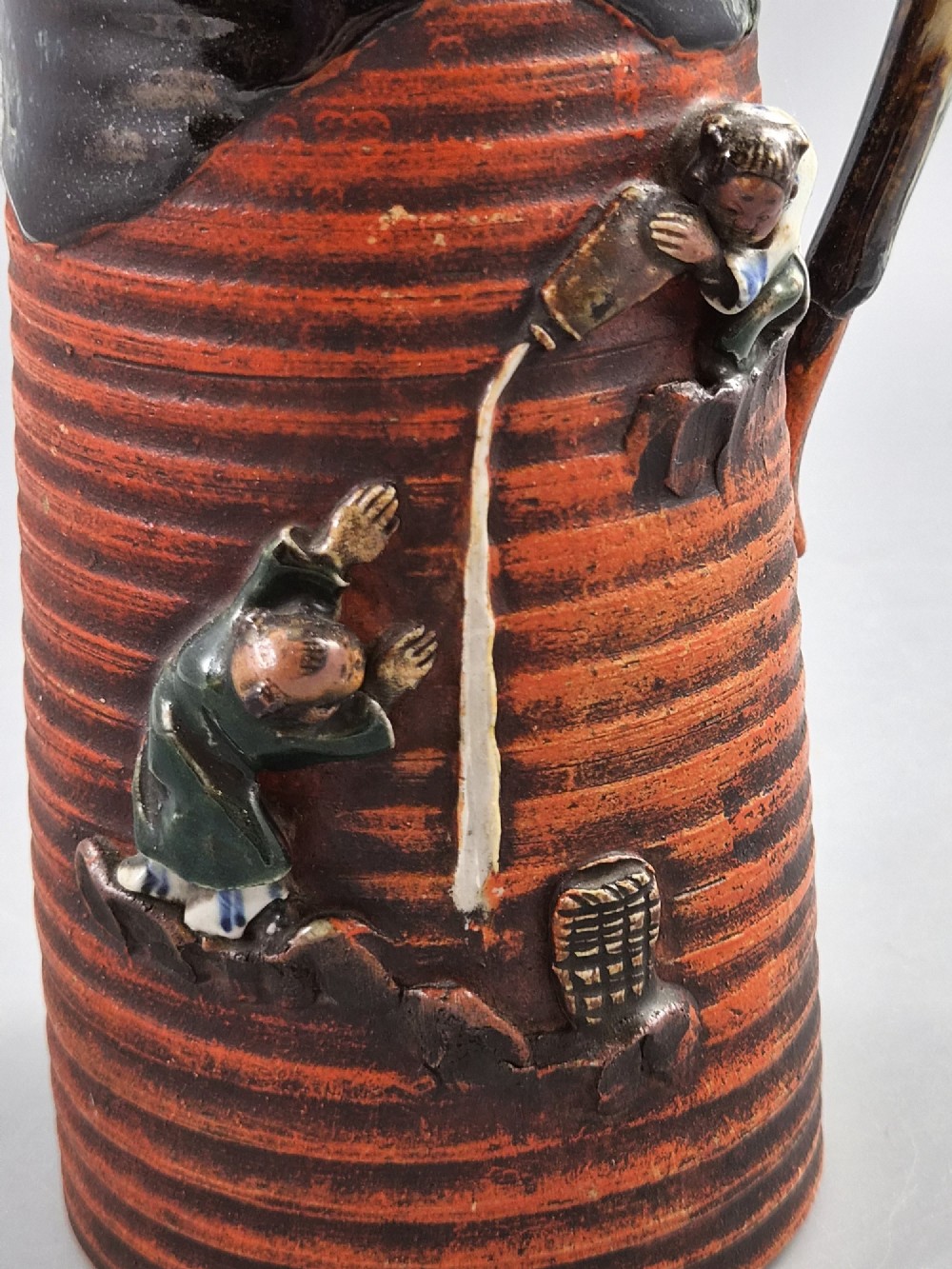 A tall Japanese Sumida Gawa ceramic pitcher featuring a ribbed terracotta body with relief figures pouring sake and a dark drip glaze near the lip. View VII
