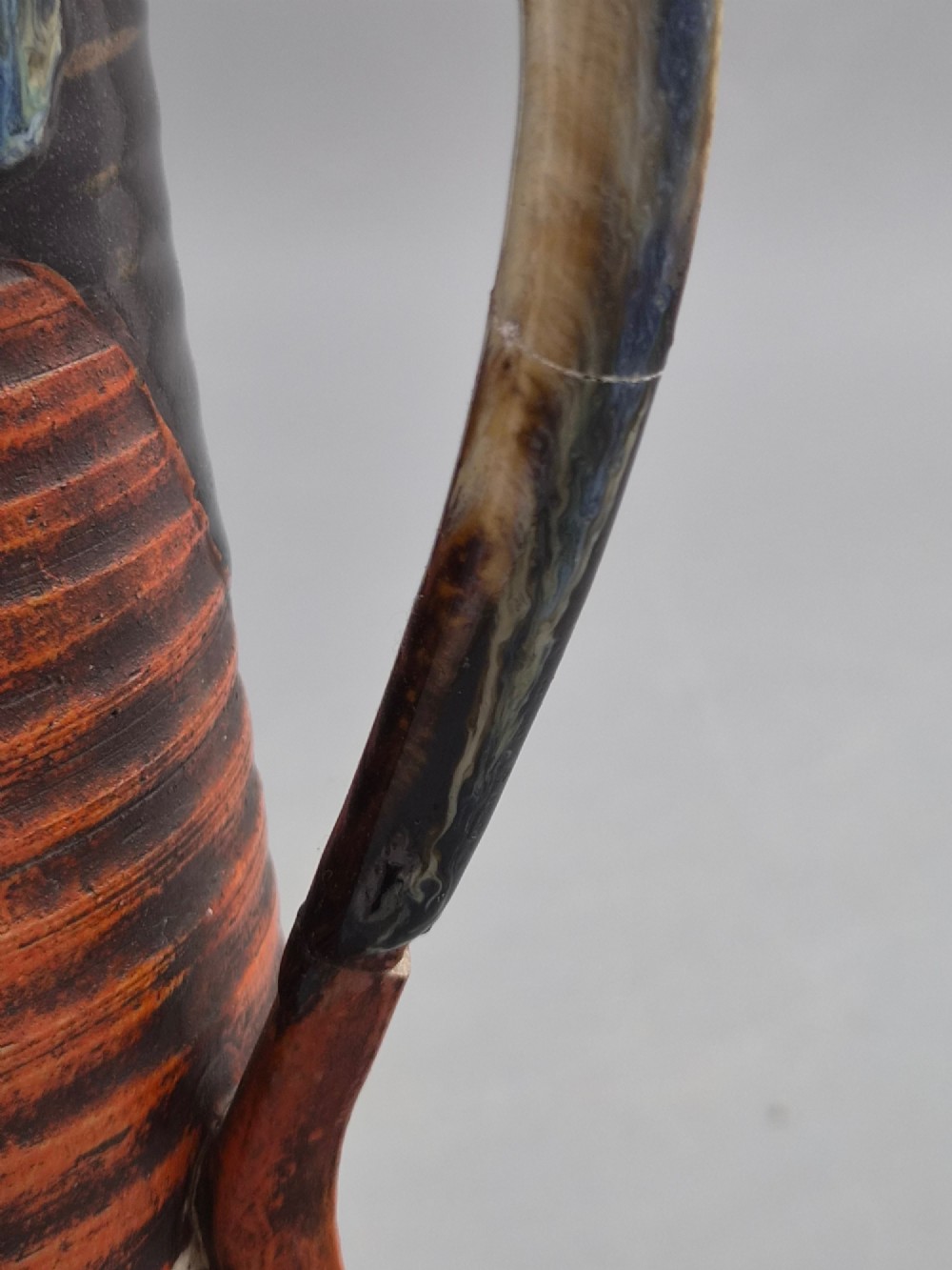 A tall Japanese Sumida Gawa ceramic pitcher featuring a ribbed terracotta body with relief figures pouring sake and a dark drip glaze near the lip. View VIII