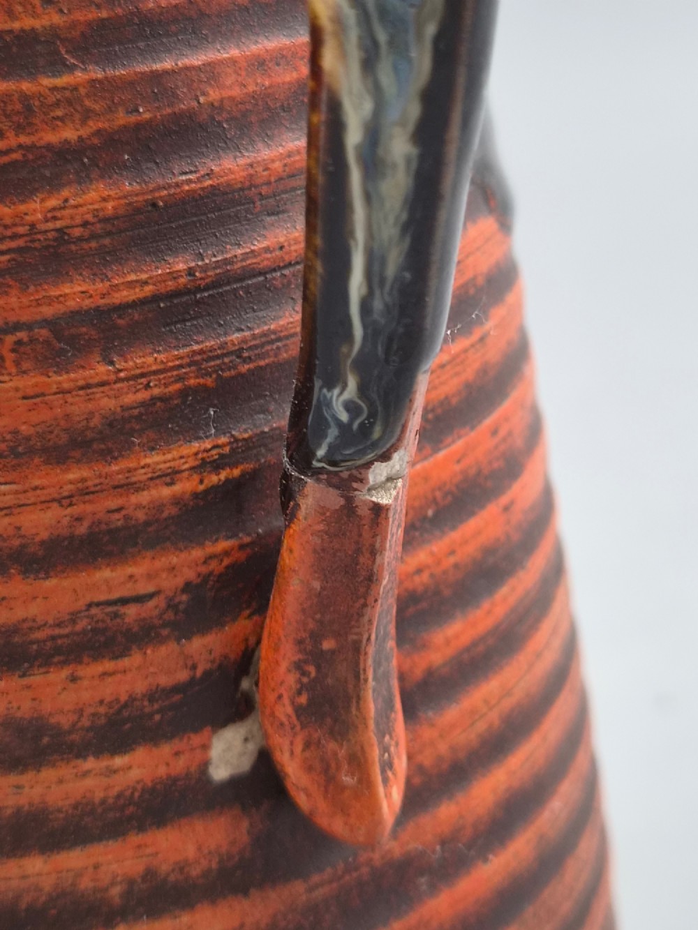 A tall Japanese Sumida Gawa ceramic pitcher featuring a ribbed terracotta body with relief figures pouring sake and a dark drip glaze near the lip. View IX