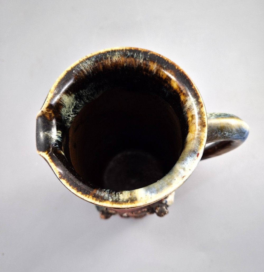 A tall Japanese Sumida Gawa ceramic pitcher featuring a ribbed terracotta body with relief figures pouring sake and a dark drip glaze near the lip. View XI