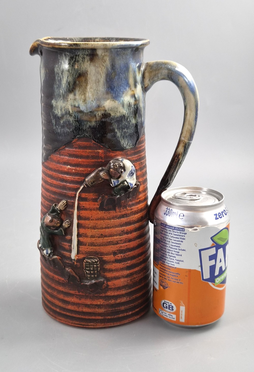 A tall Japanese Sumida Gawa ceramic pitcher featuring a ribbed terracotta body with relief figures pouring sake and a dark drip glaze near the lip. View XIII