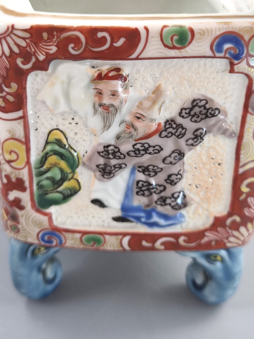 A square Japanese Kutani porcelain incense burner featuring a karashishi lion finial on the lid, hand-painted figural panels, and blue elephant-head legs. View X