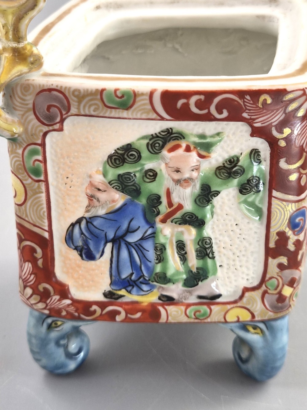 A square Japanese Kutani porcelain incense burner featuring a karashishi lion finial on the lid, hand-painted figural panels, and blue elephant-head legs. View XI