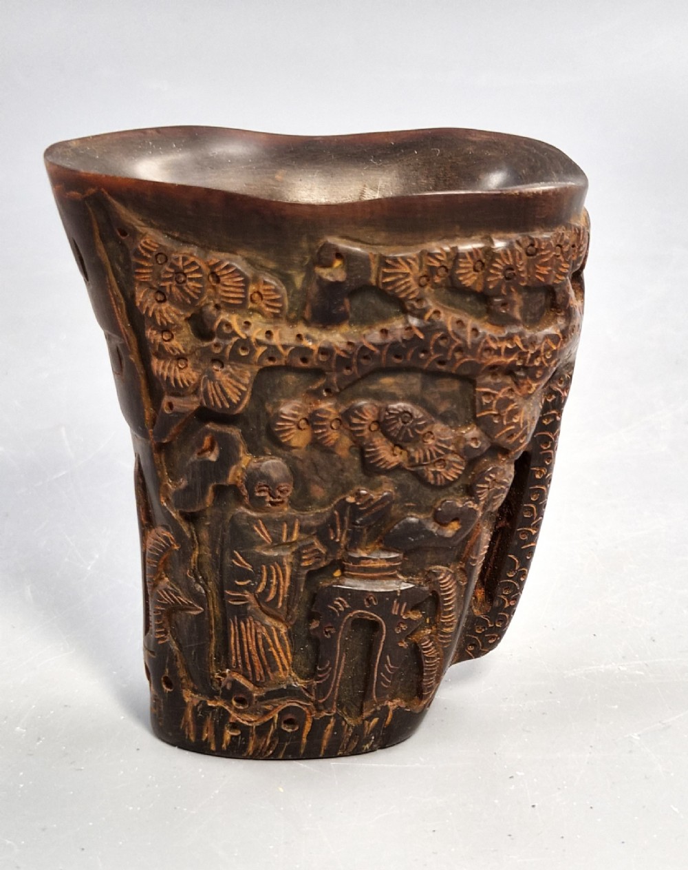 A dark brown carved water buffalo horn libation cup featuring relief carvings of immortals standing under pine trees.