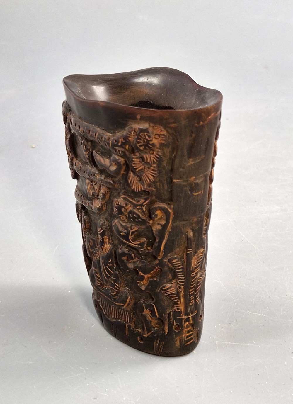 A dark brown carved water buffalo horn libation cup featuring relief carvings of immortals standing under pine trees. View V