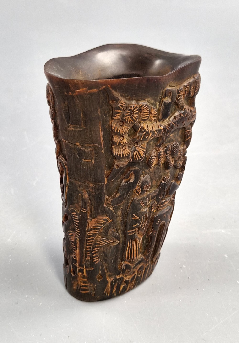A dark brown carved water buffalo horn libation cup featuring relief carvings of immortals standing under pine trees. View VI
