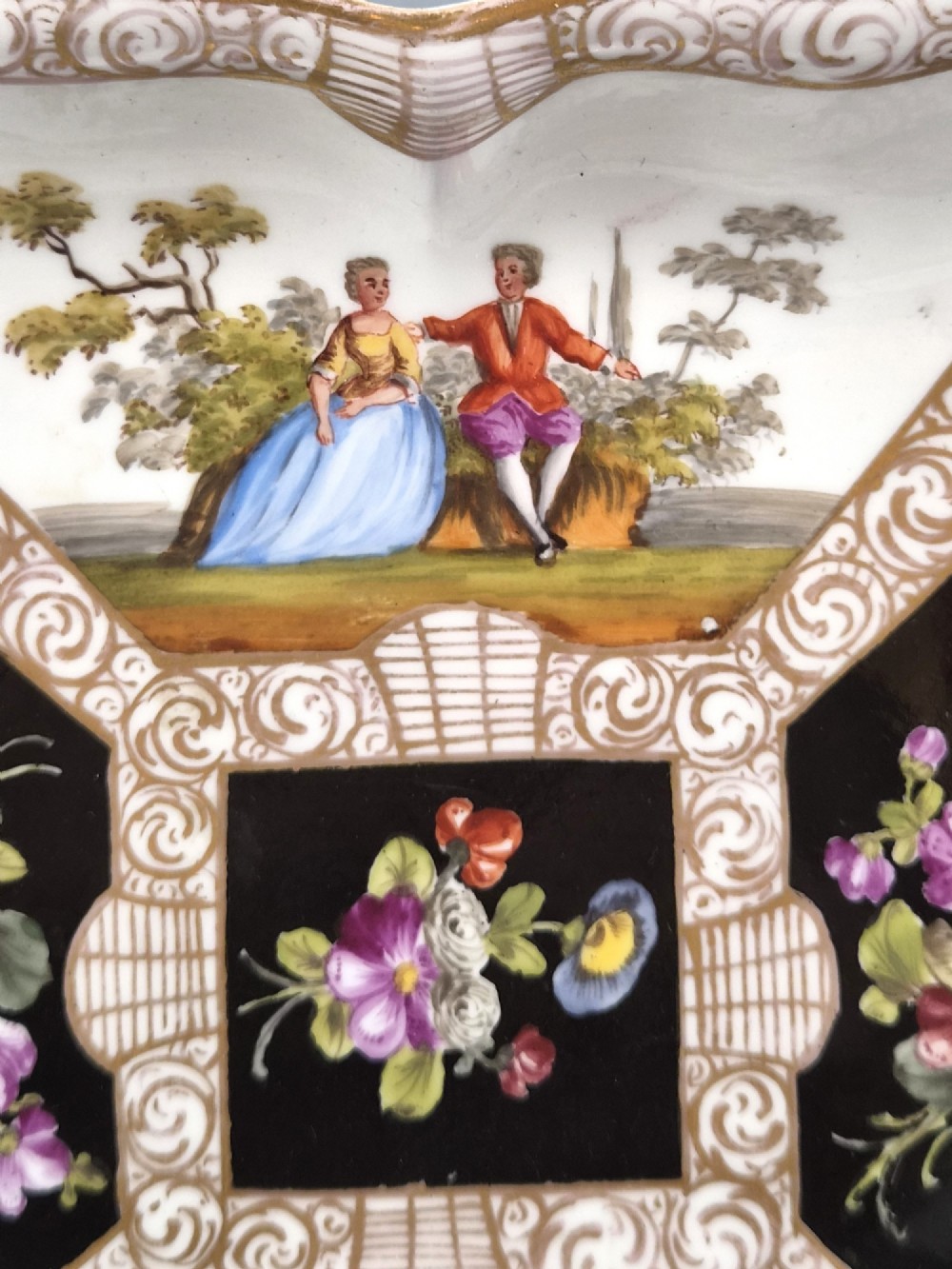A square shaped Meissen porcelain dish decorated with hand-painted Watteau scenes, floral reserves on a black ground, and gilded scroll borders. View II