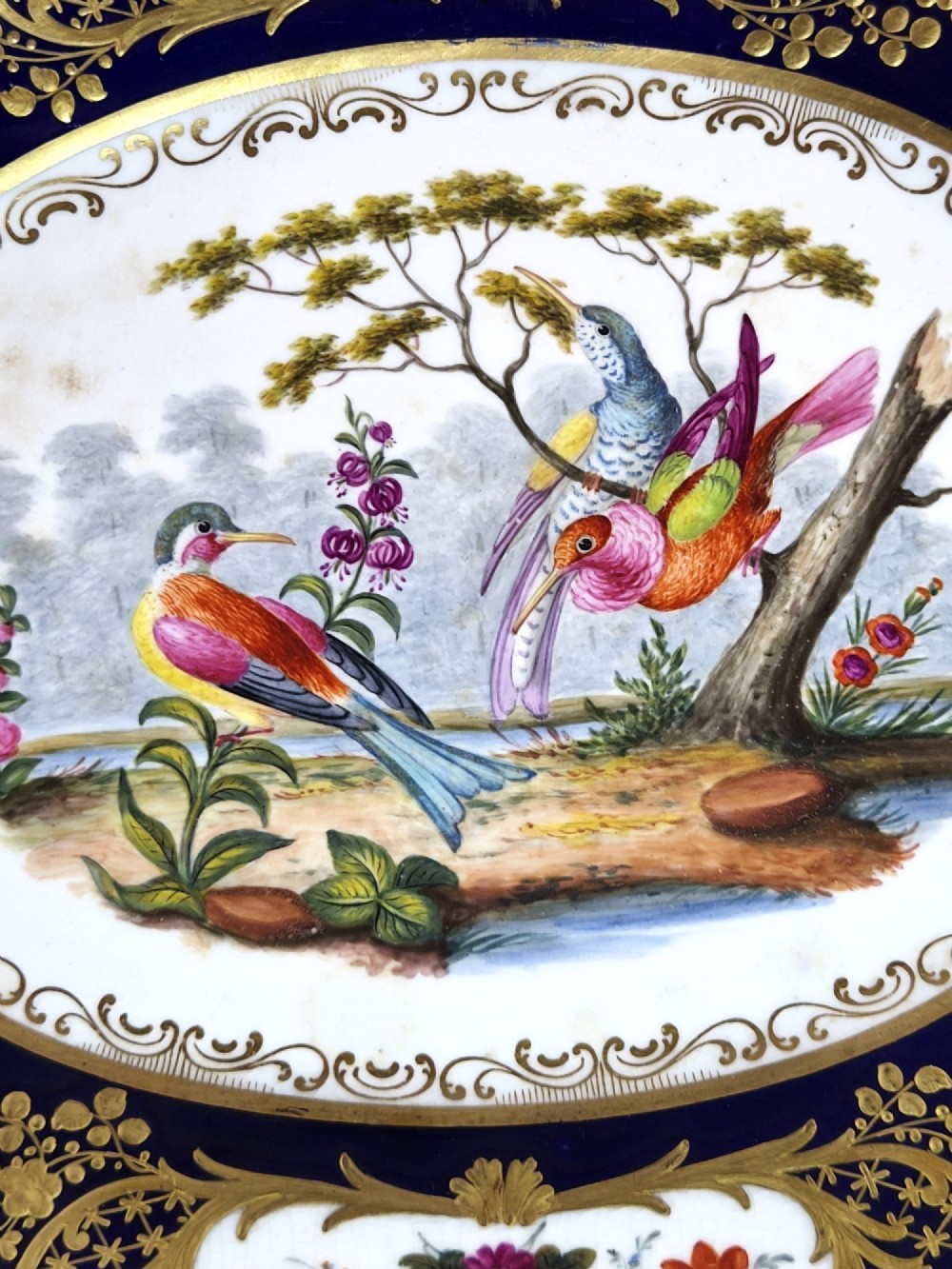 An oval porcelain dish featuring a dark cobalt blue rim with gold scrollwork, reticulated handles, floral panels, and a central hand-painted scene of colorful birds perched on branches. View II