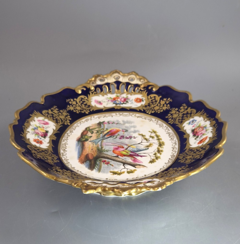 An oval porcelain dish featuring a dark cobalt blue rim with gold scrollwork, reticulated handles, floral panels, and a central hand-painted scene of colorful birds perched on branches. View VI