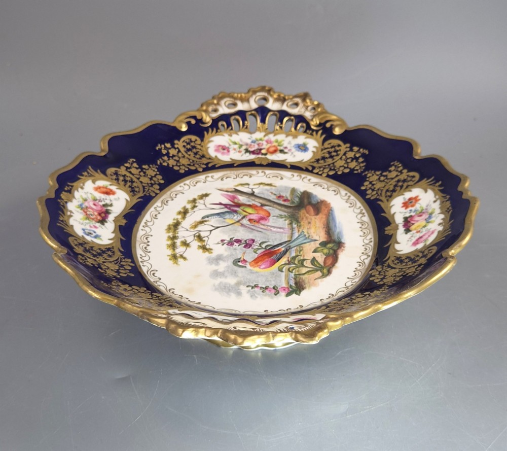 An oval porcelain dish featuring a dark cobalt blue rim with gold scrollwork, reticulated handles, floral panels, and a central hand-painted scene of colorful birds perched on branches. View VIII