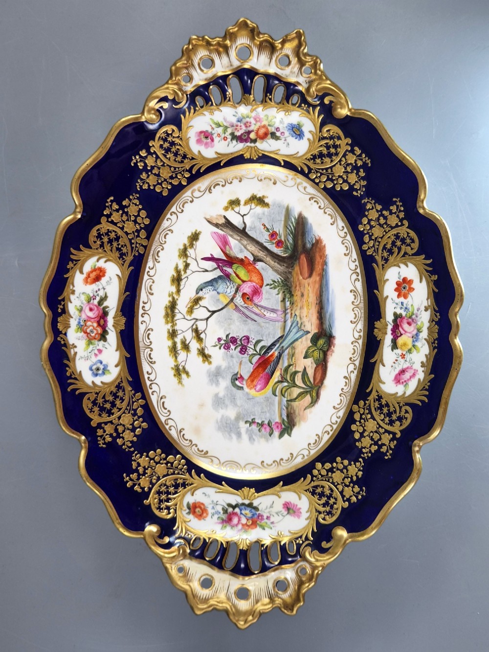 An oval porcelain dish featuring a dark cobalt blue rim with gold scrollwork, reticulated handles, floral panels, and a central hand-painted scene of colorful birds perched on branches. View IX