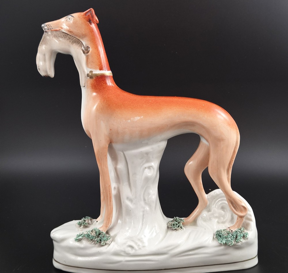 A pair of antique Victorian Staffordshire earthenware figures depicting standing orange greyhounds holding grey hares in their jaws on white bases. View II