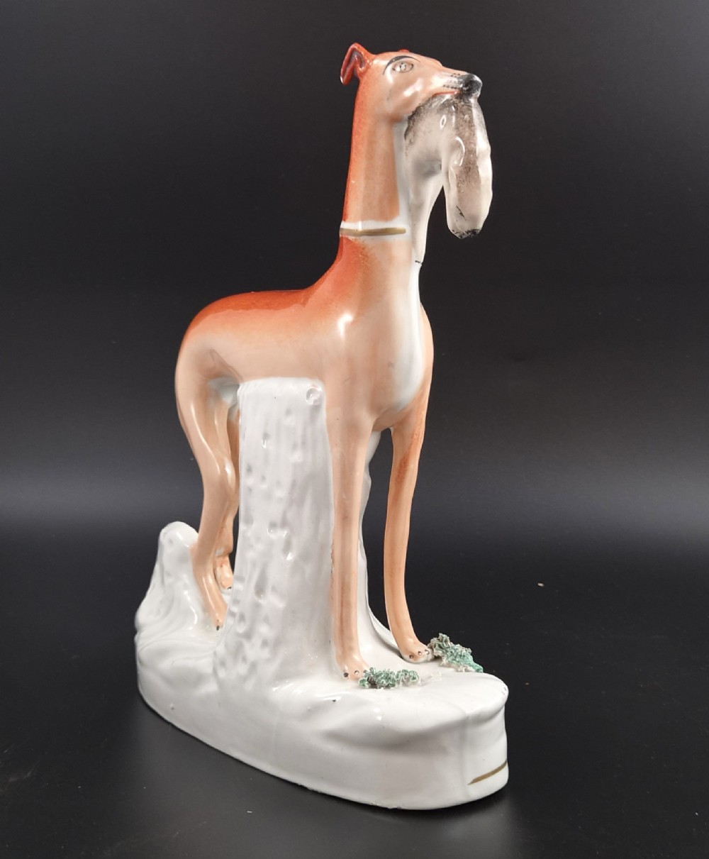 A pair of antique Victorian Staffordshire earthenware figures depicting standing orange greyhounds holding grey hares in their jaws on white bases. View V