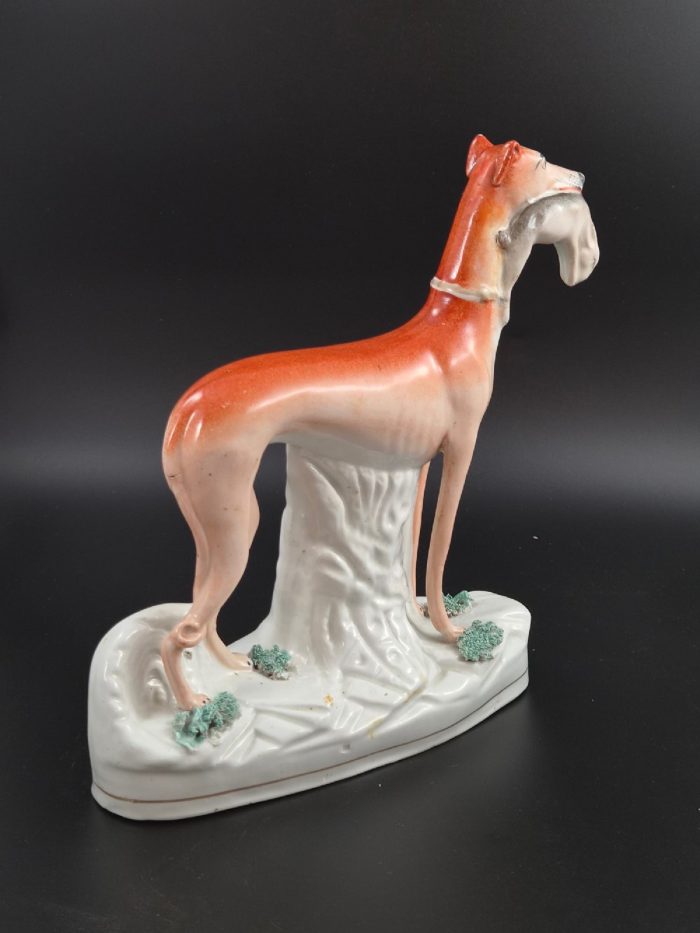 A pair of antique Victorian Staffordshire earthenware figures depicting standing orange greyhounds holding grey hares in their jaws on white bases. View IX