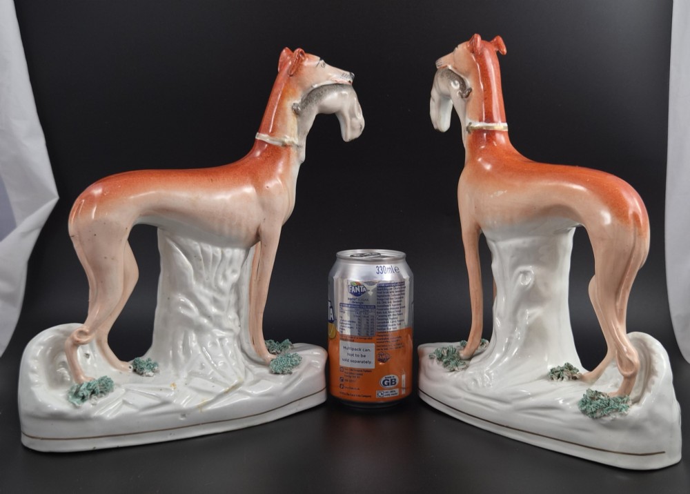 A pair of antique Victorian Staffordshire earthenware figures depicting standing orange greyhounds holding grey hares in their jaws on white bases. View XV