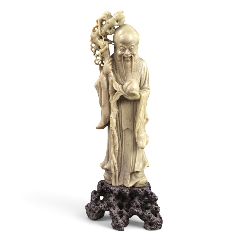 A late 19th-century carved pale soapstone figure of Shoulao holding a peach and staff, standing on a dark stone pierced rockwork base.