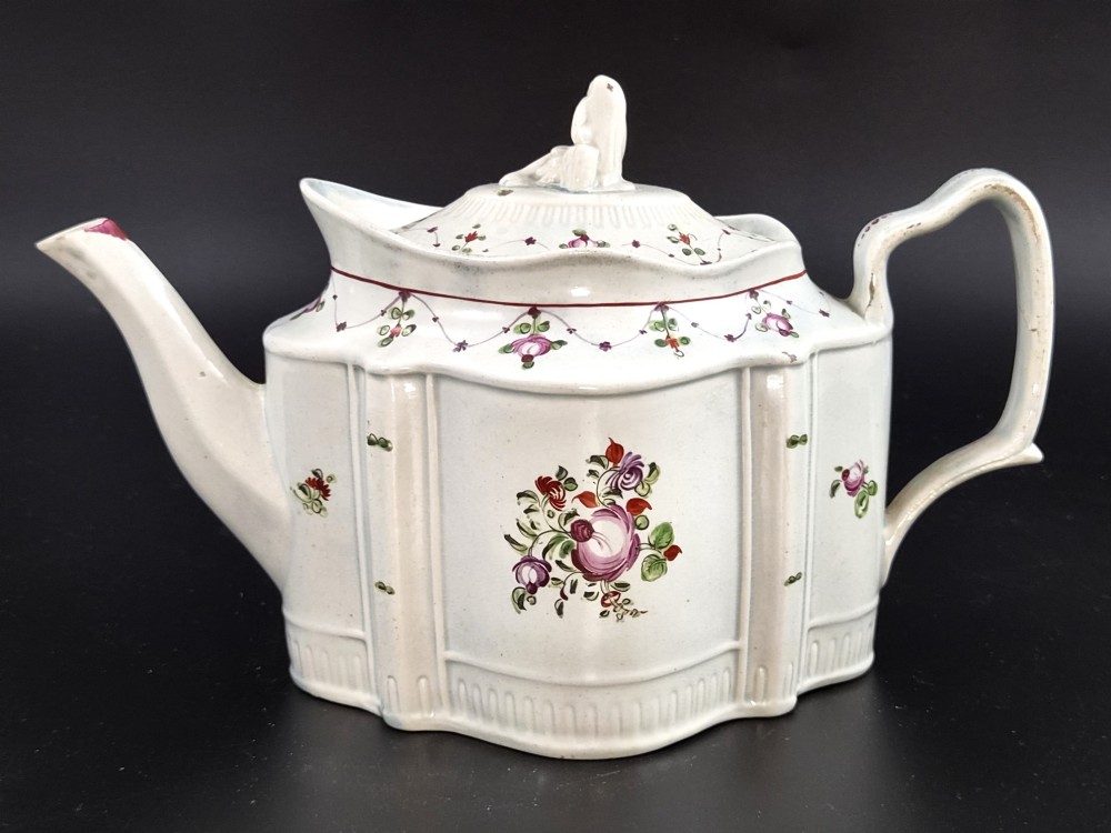An off-white earthenware commode-shaped teapot decorated with hand-painted floral sprays, red borders, and a seated widow finial on the lid.
