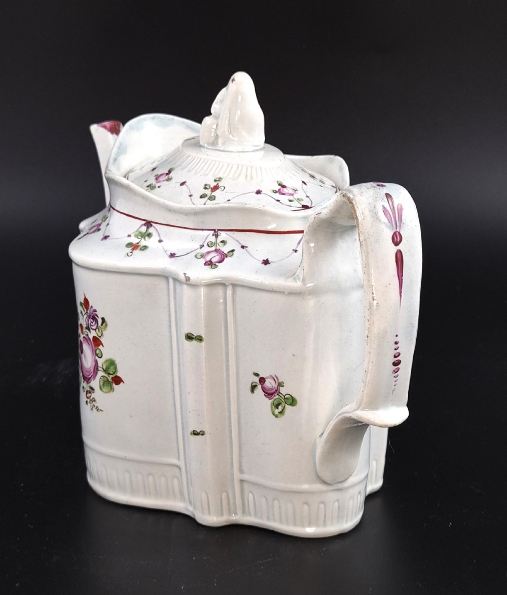 An off-white earthenware commode-shaped teapot decorated with hand-painted floral sprays, red borders, and a seated widow finial on the lid. View II