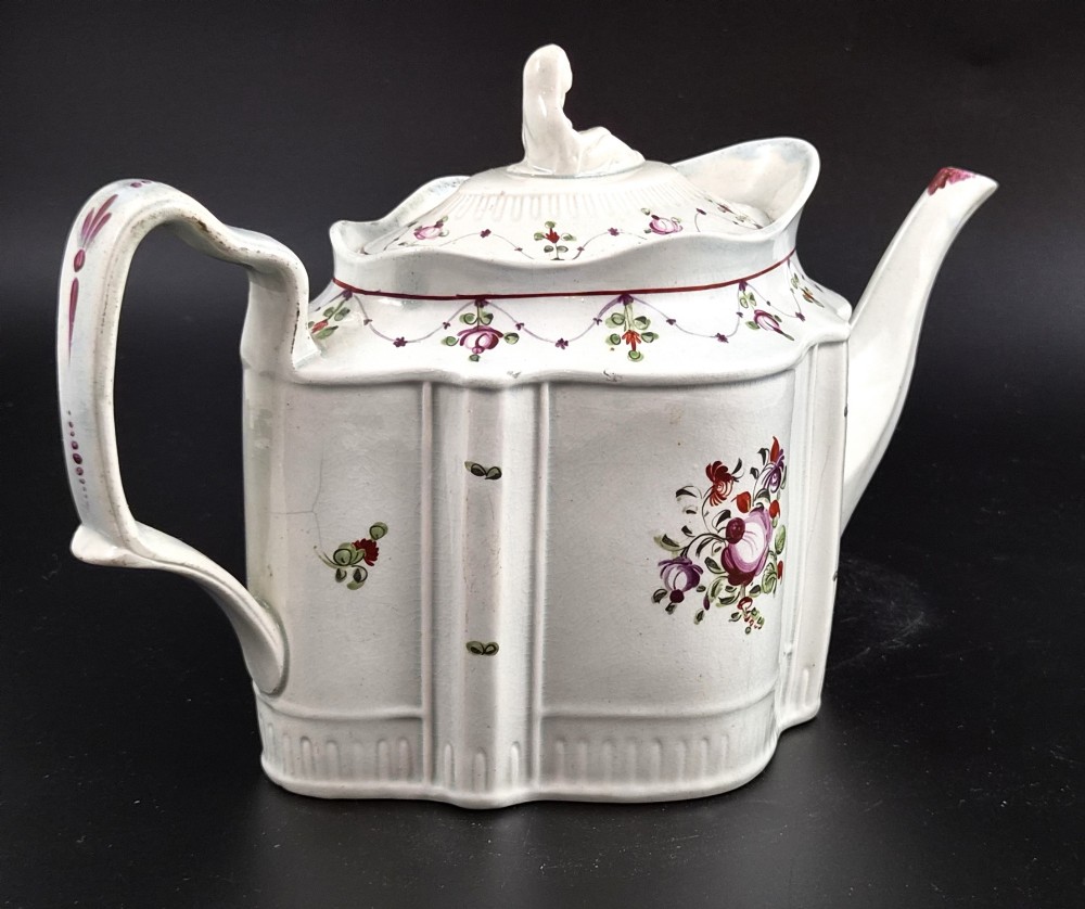 An off-white earthenware commode-shaped teapot decorated with hand-painted floral sprays, red borders, and a seated widow finial on the lid. View III