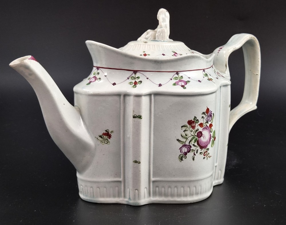 An off-white earthenware commode-shaped teapot decorated with hand-painted floral sprays, red borders, and a seated widow finial on the lid. View V