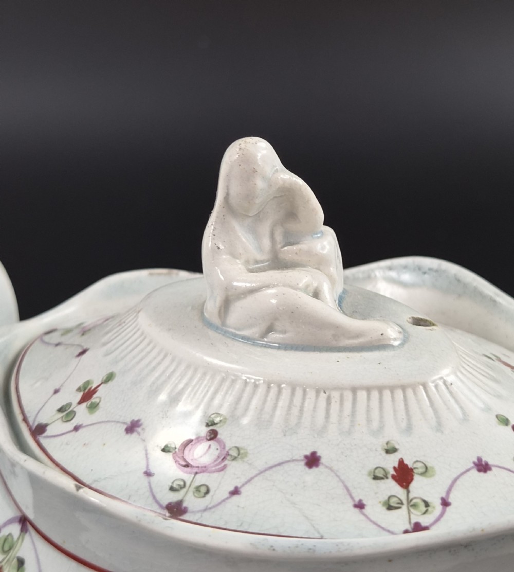 An off-white earthenware commode-shaped teapot decorated with hand-painted floral sprays, red borders, and a seated widow finial on the lid. View VI