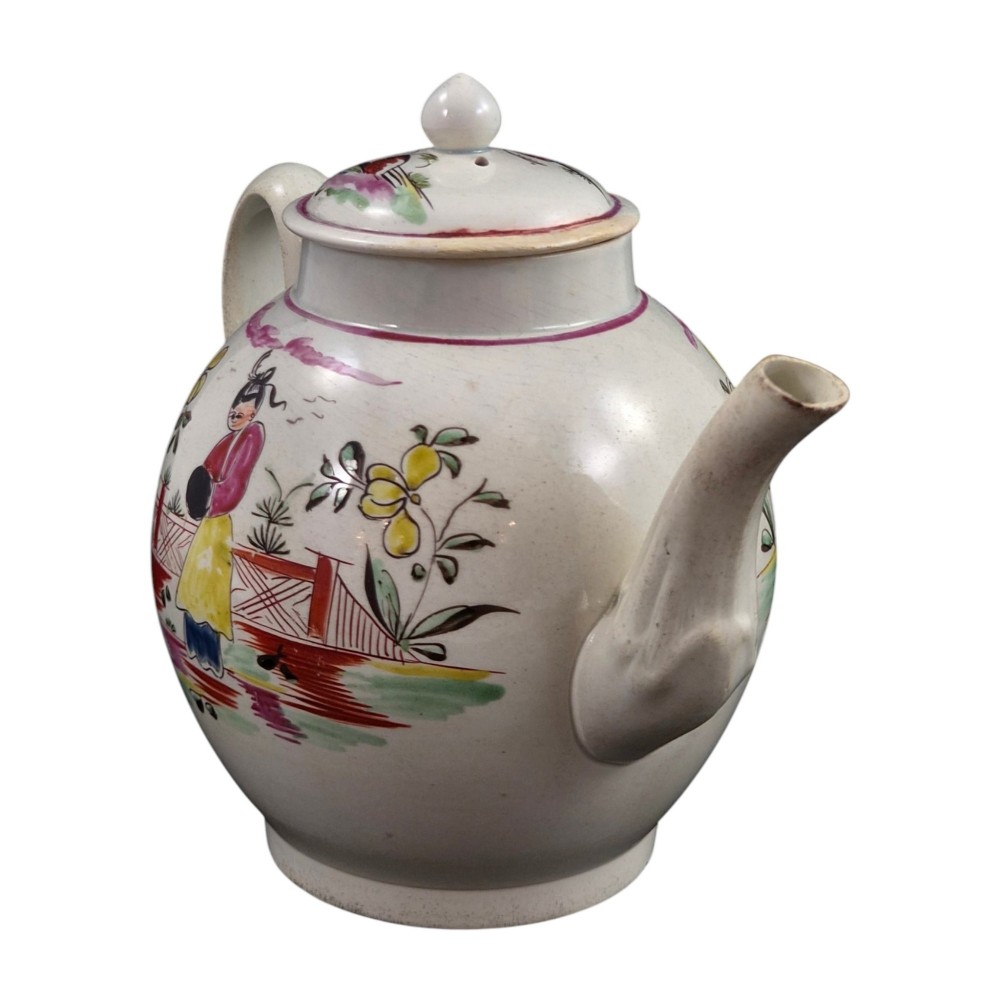 An 18th-century creamware teapot featuring hand-painted polychrome Chinoiserie figures on a cream glaze background with a curved spout and loop handle. View V
