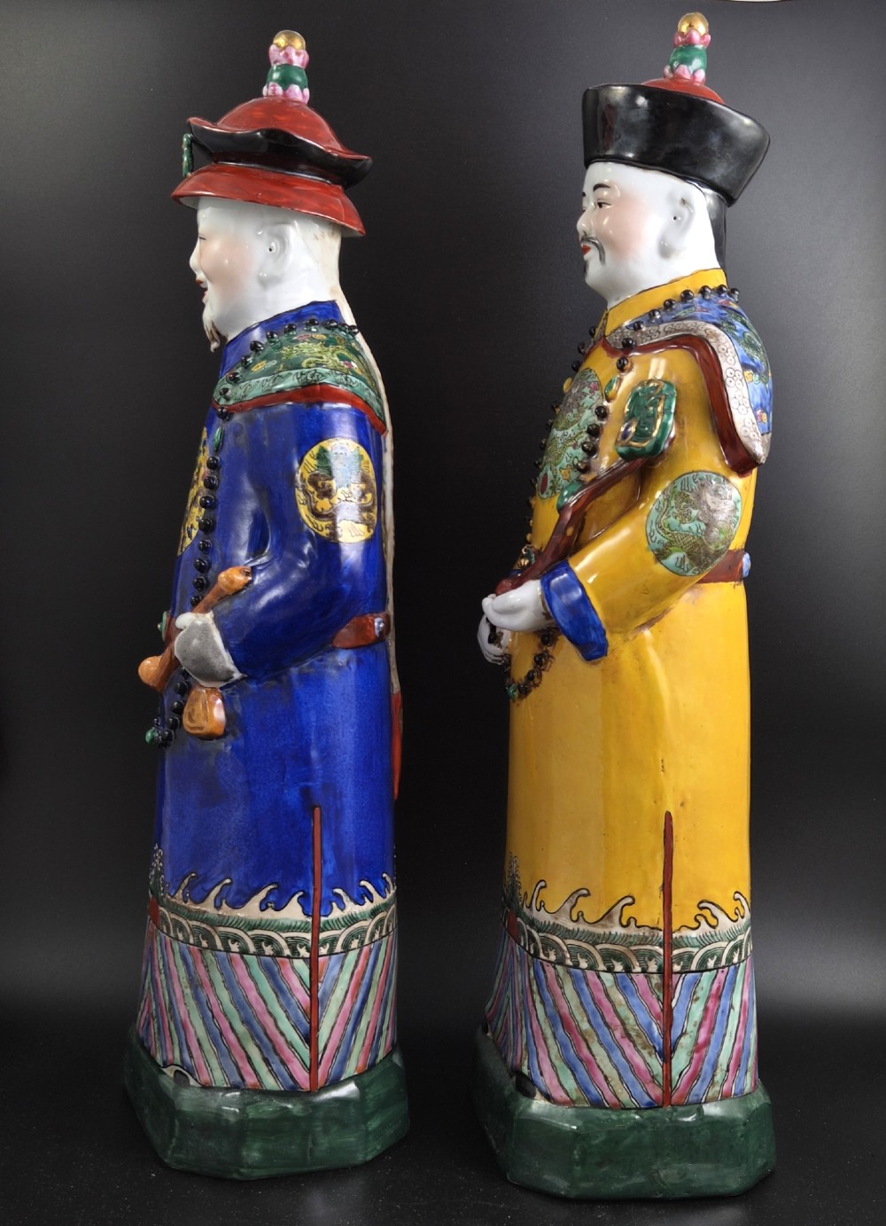 A pair of tall Chinese porcelain figures wearing traditional Qing Dynasty imperial robes in bright yellow and royal blue, adorned with dragon roundels and court beads. View II