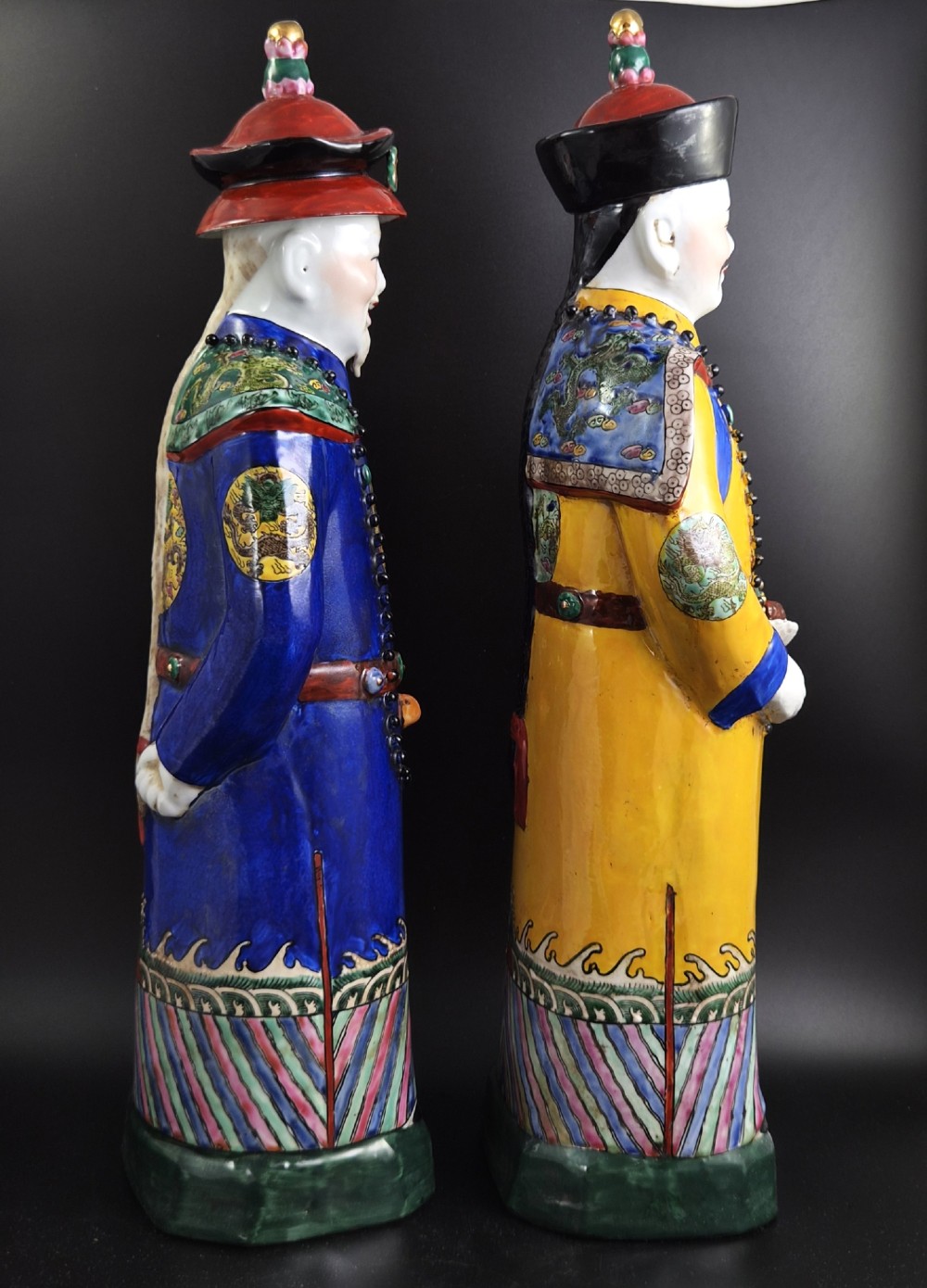 A pair of tall Chinese porcelain figures wearing traditional Qing Dynasty imperial robes in bright yellow and royal blue, adorned with dragon roundels and court beads. View IV