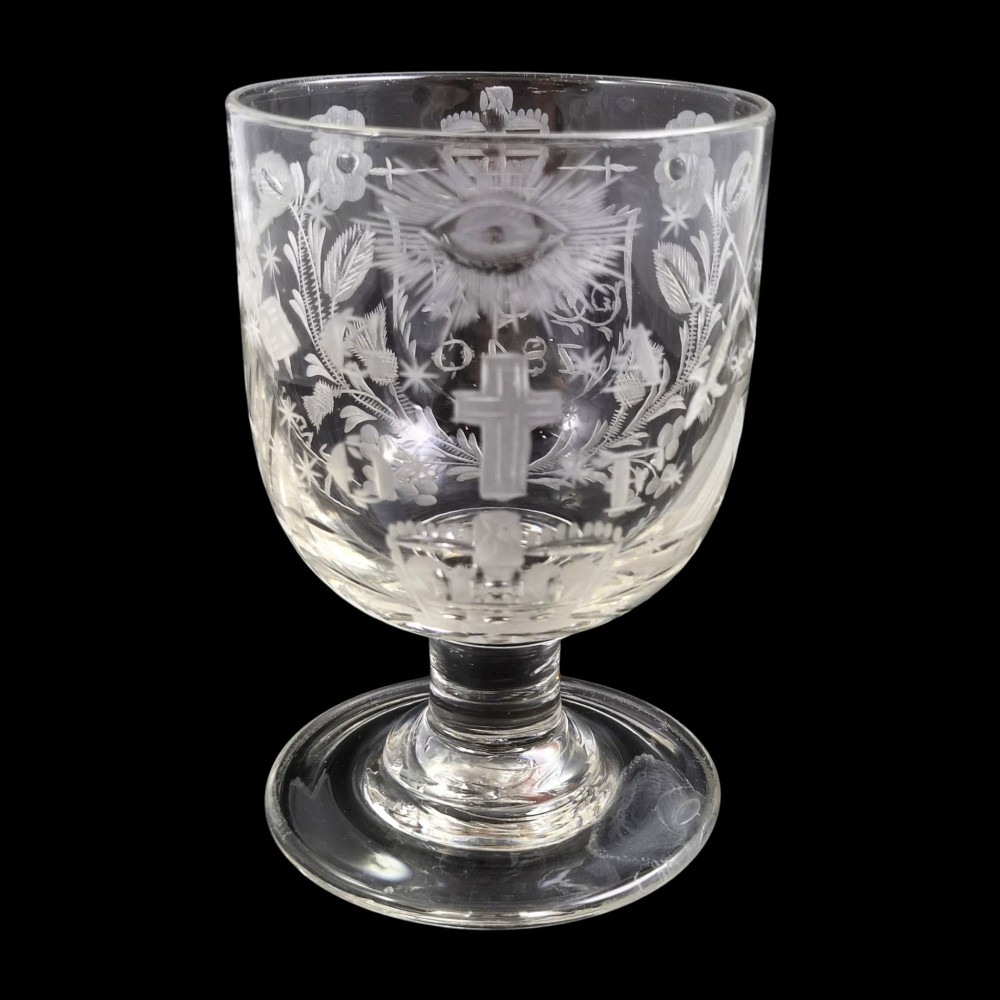 A clear lead-glass rummer featuring detailed wheel-engraved fraternal symbols, including a central cross, all-seeing eye, crowns, and floral garlands, resting on a short stem and circular foot.
