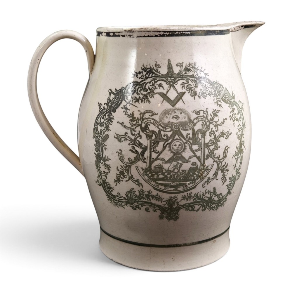 A Georgian Staffordshire creamware jug featuring transfer-printed olive-green Masonic iconography framed by a leafy scrollwork wreath.