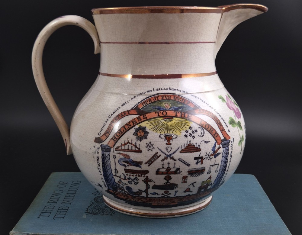 A cream earthenware jug featuring hand-painted floral motifs, copper lustre trim, and a printed Orange Order emblematic coat of arms with religious and fraternal symbols.