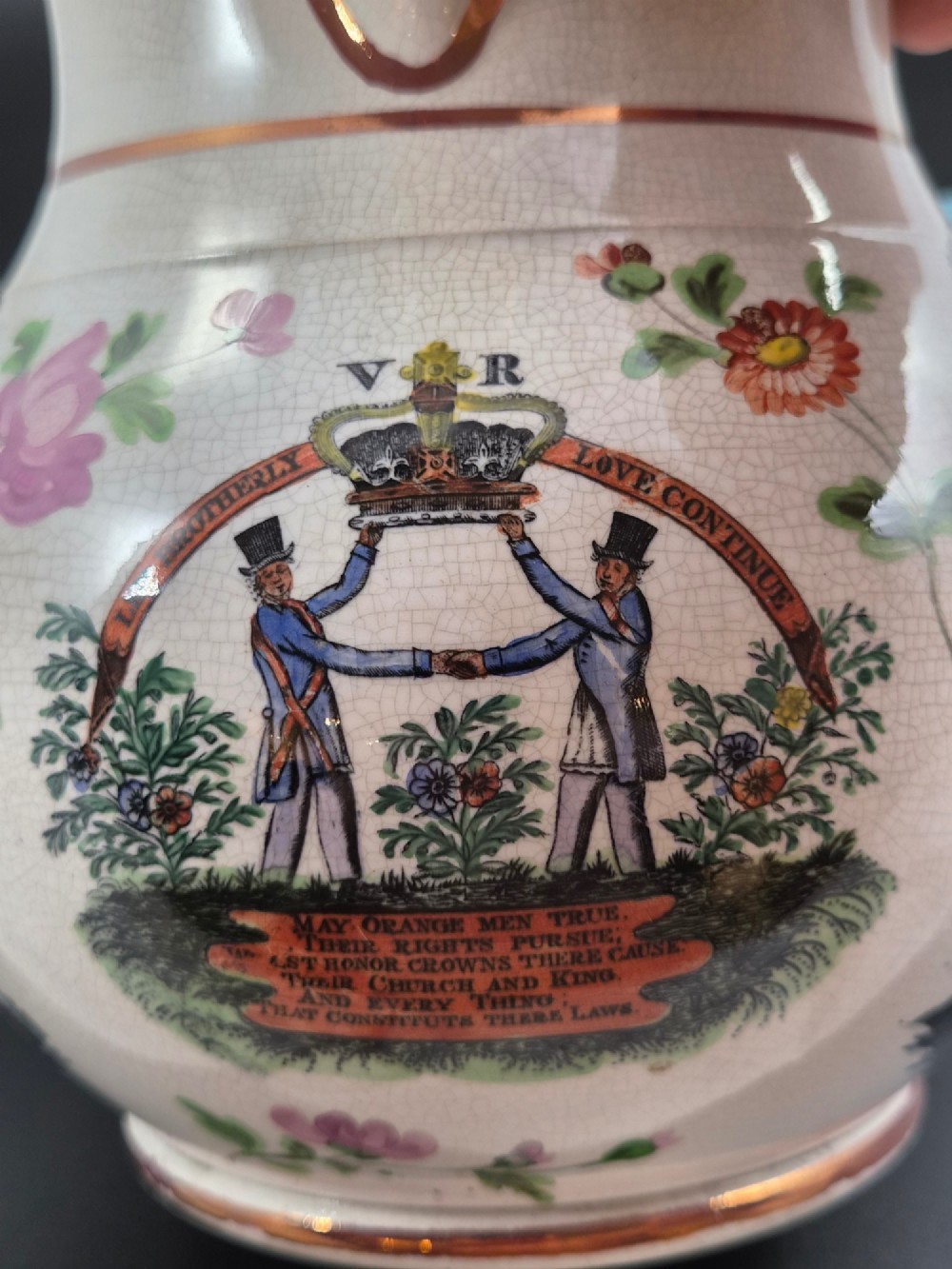 A cream earthenware jug featuring hand-painted floral motifs, copper lustre trim, and a printed Orange Order emblematic coat of arms with religious and fraternal symbols. View X