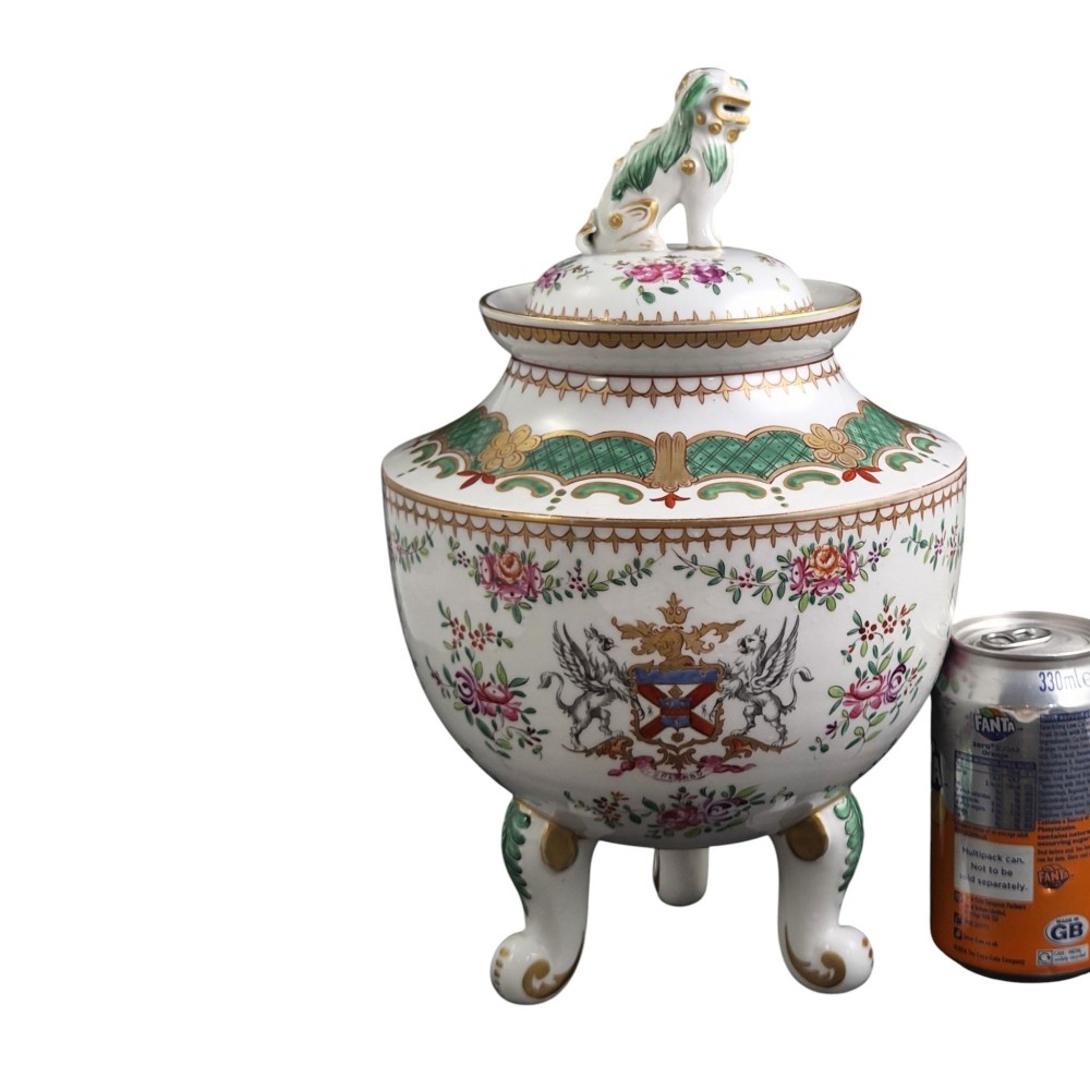A white hard-paste porcelain covered jar on three cabriole legs, decorated with floral enamel sprays, an armorial crest, green lattice borders, and topped with a foo dog lid finial. View IX
