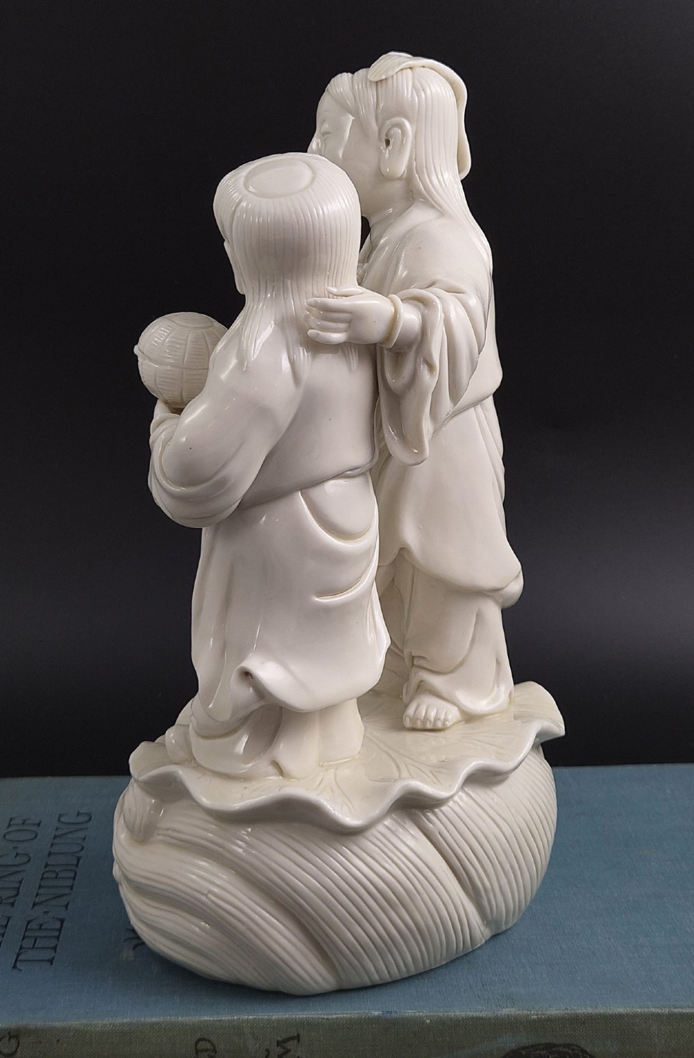 A Qing Dynasty Blanc-de-Chine white porcelain figure group depicting the Hehe Erxian standing on a stylised lotus and wave base. View V