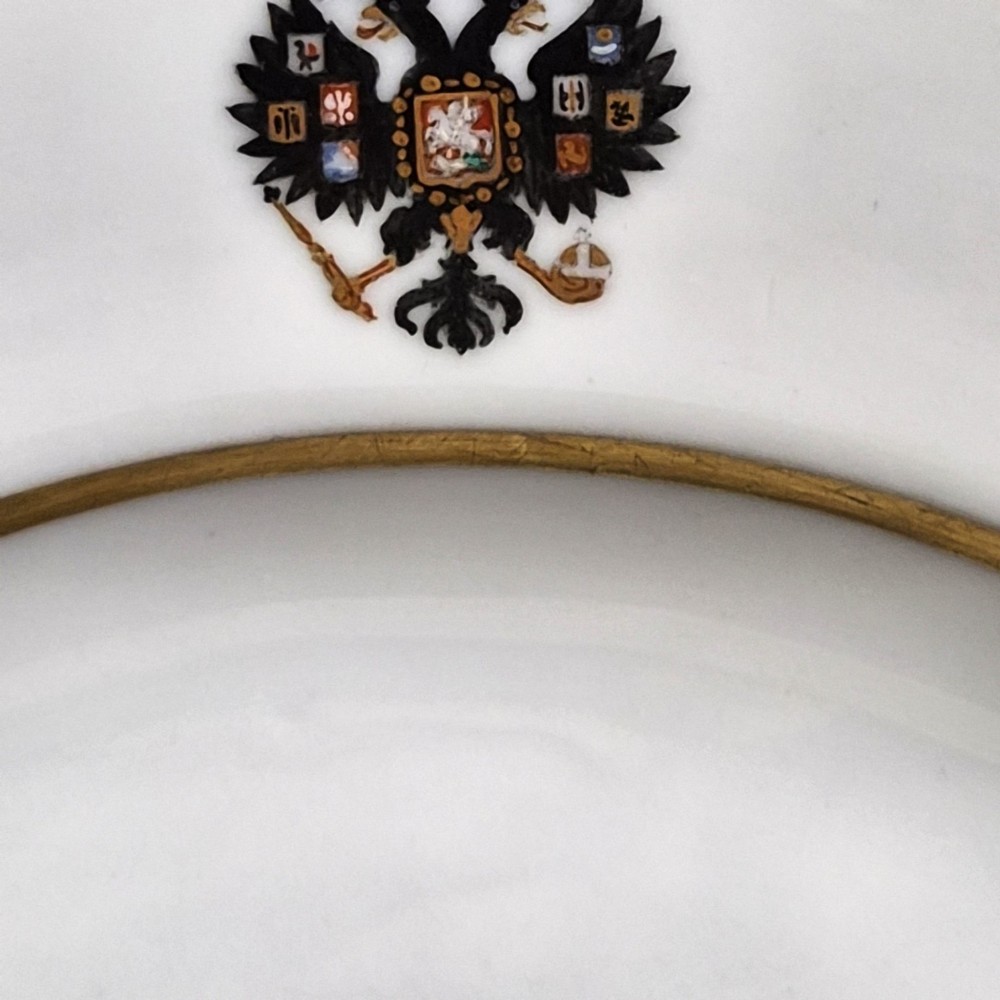 A circular white porcelain plate with twin gilded rim borders and a small Russian Imperial double-headed eagle crest near the top edge. View V