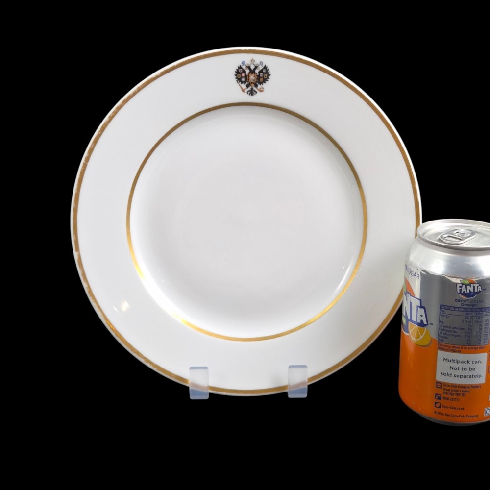 A circular white porcelain plate with twin gilded rim borders and a small Russian Imperial double-headed eagle crest near the top edge. View VI