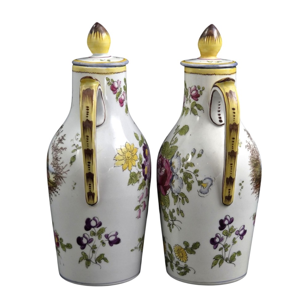 A pair of 18th-century white tin-glazed earthenware oil and vinegar jugs decorated with hand-painted floral motifs and fitted with yellow finial covers. View X