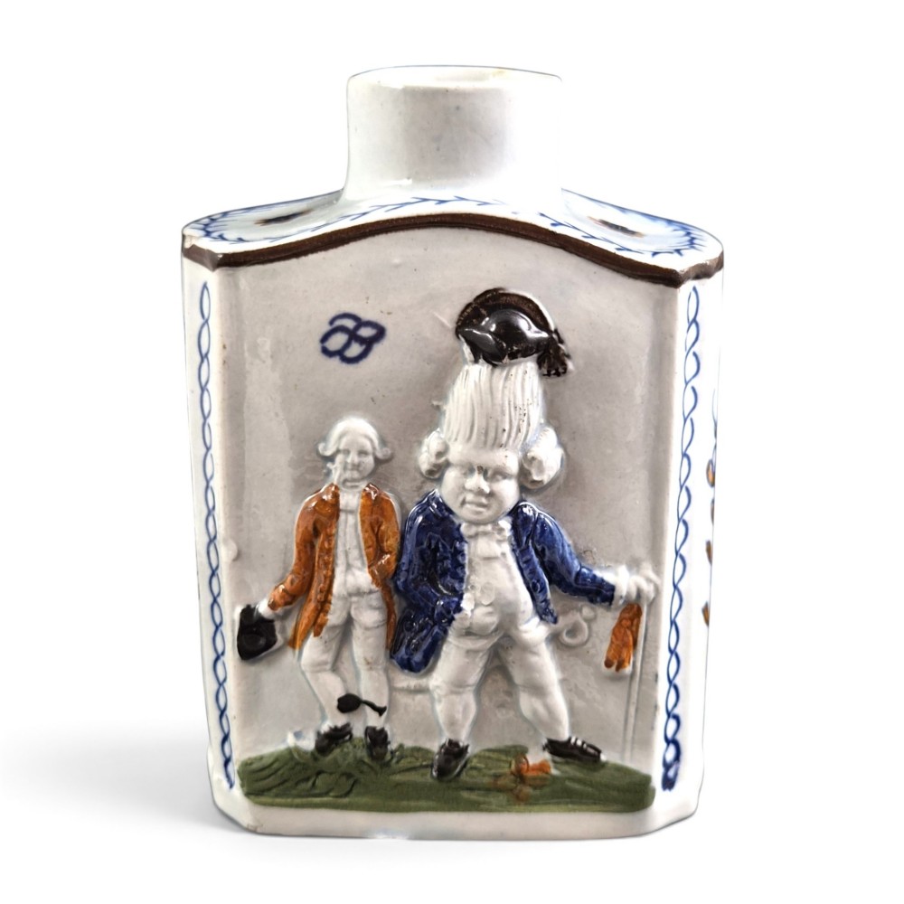 An 18th-century Prattware earthenware tea canister featuring relief-moulded depictions of two Georgian Macaroni figures in blue, ochre, and green underglaze glazes against an off-white background.