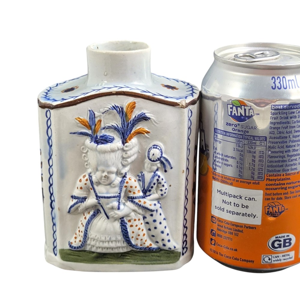 An 18th-century Prattware earthenware tea canister featuring relief-moulded depictions of two Georgian Macaroni figures in blue, ochre, and green underglaze glazes against an off-white background. View VI
